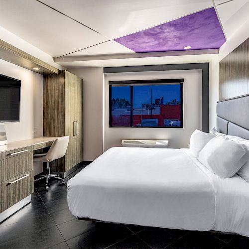 A modern hotel room with a large bed, desk area, and purple ceiling.