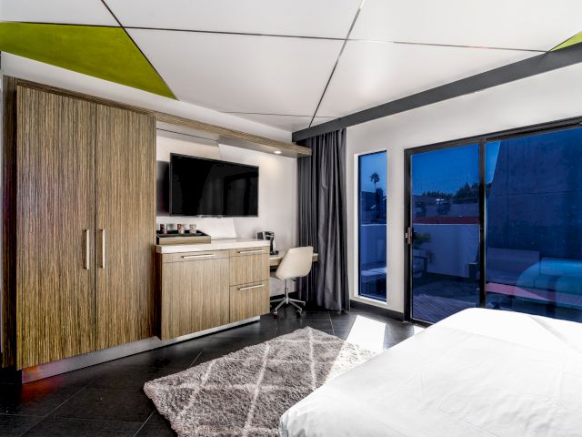 A modern hotel room with a large bed, desk area, closet, TV, and sliding glass doors leading to a patio.