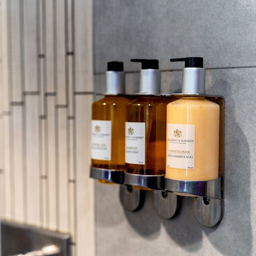 Three dispensing bottles of toiletries mounted on a gray tile wall.