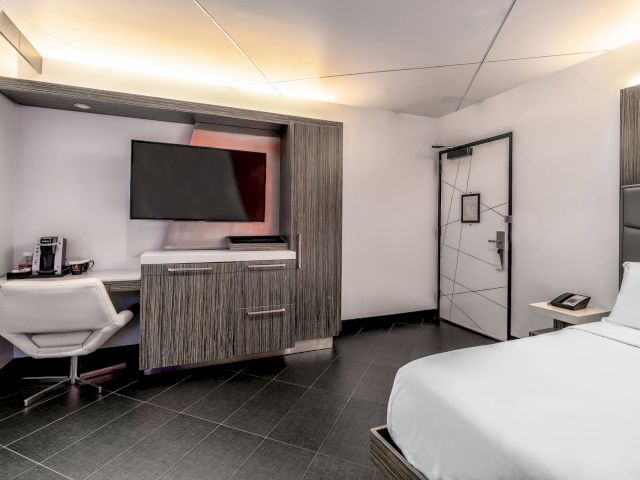 A modern hotel room with a desk, TV, bed, and chair.