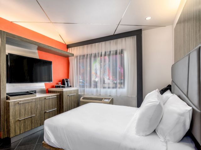 A hotel room with a large bed, flat-screen TV, and window with sheer curtains.