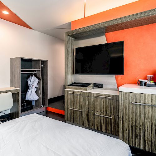 A hotel room with a large bed, a flat-screen TV mounted on an orange accent wall, and a built-in closet.