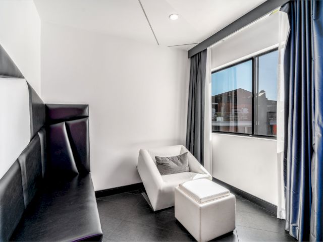 A modern hotel room with a black leather bed, white armchair, and ottoman, facing a city view.