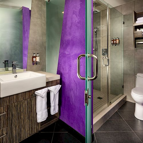 A modern bathroom with a glass-enclosed shower, a wooden vanity with a white sink, and a purple accent wall.