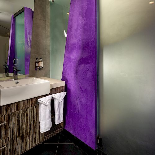 A modern bathroom sink area with a purple accent wall, brown wood cabinets, and white sink.