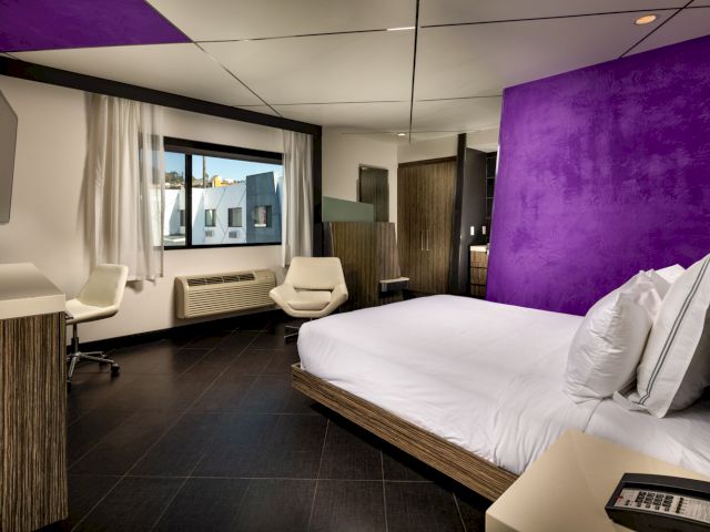 A modern hotel room with a purple accent wall, a large bed, and a desk area.