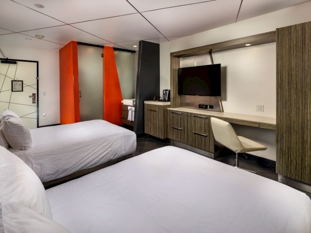 A wide view of a modern hotel room with two twin beds, a desk with a TV, and a bathroom door.
