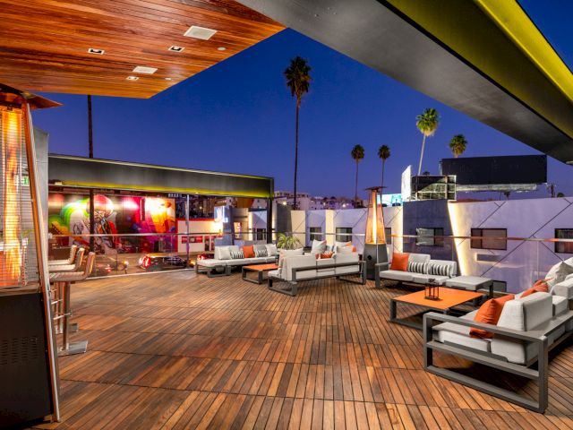 A rooftop patio with a wooden deck, seating areas, and palm trees in the background at dusk.
