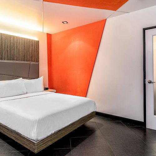 A modern hotel room with a white bed, orange accent wall, and dark floor.