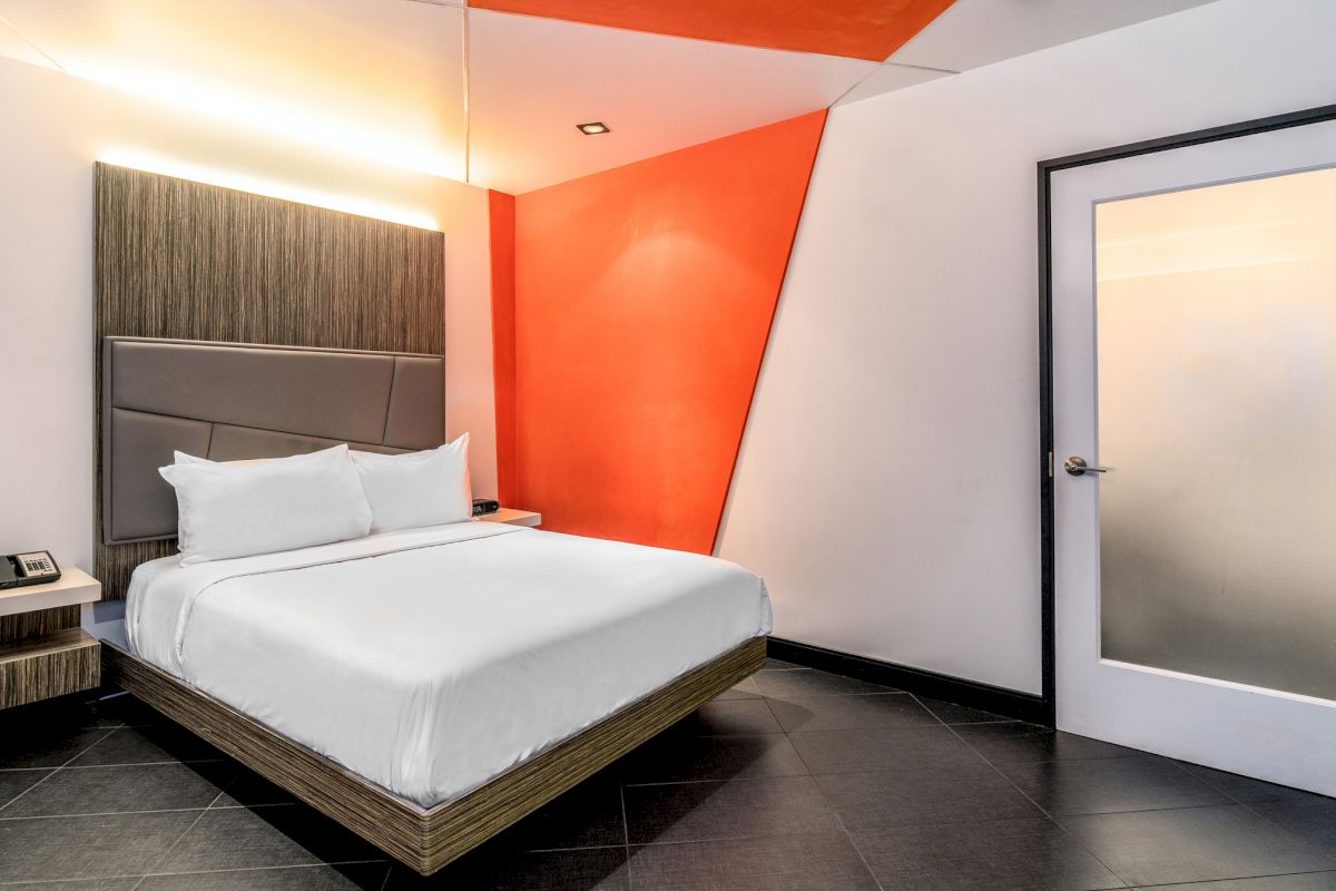 A modern hotel room with a platform bed, floating headboard, and orange accent wall.