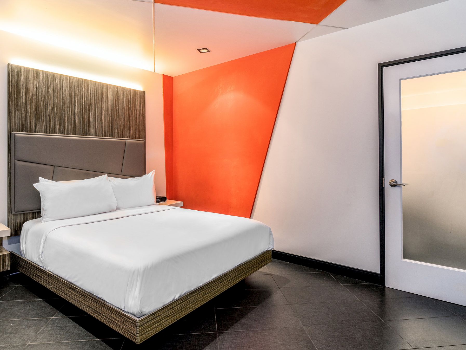 A modern hotel room with a white bed, orange accent wall, and dark floor.