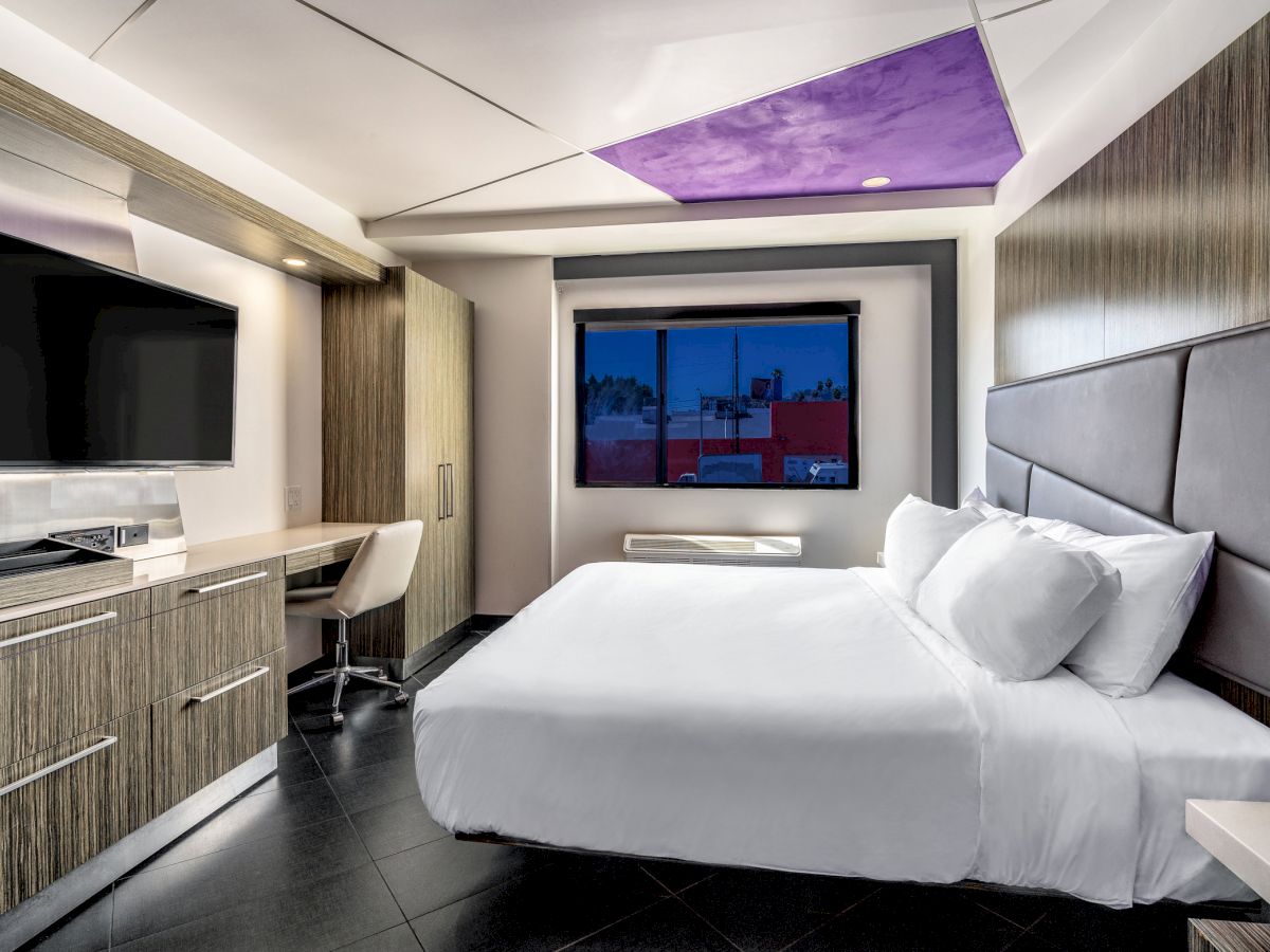 A modern hotel room with a large bed, desk, TV, and window.