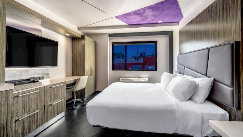 A modern hotel room with a large bed, desk, TV, and purple ceiling.