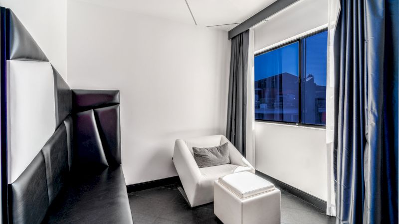 A hotel room with a black bed, white chair, and window with gray curtains.