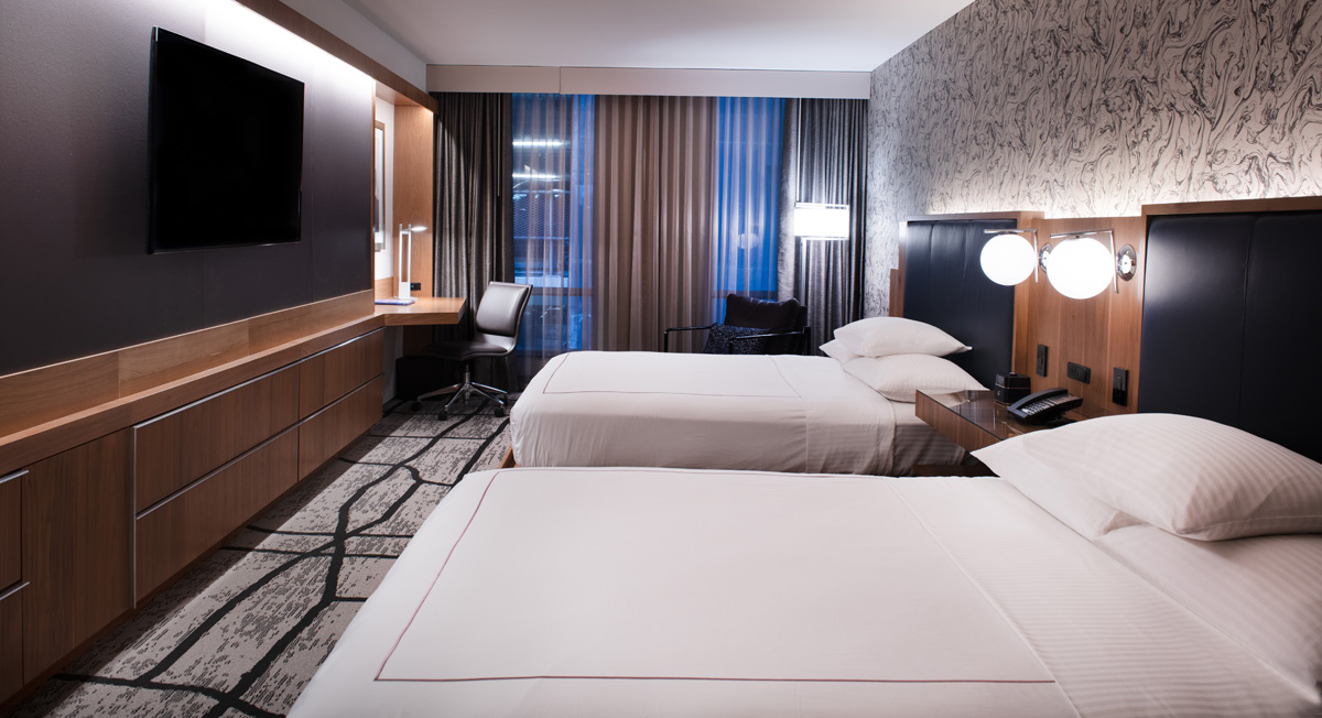 A hotel room with two beds, a desk, a TV, and a window.